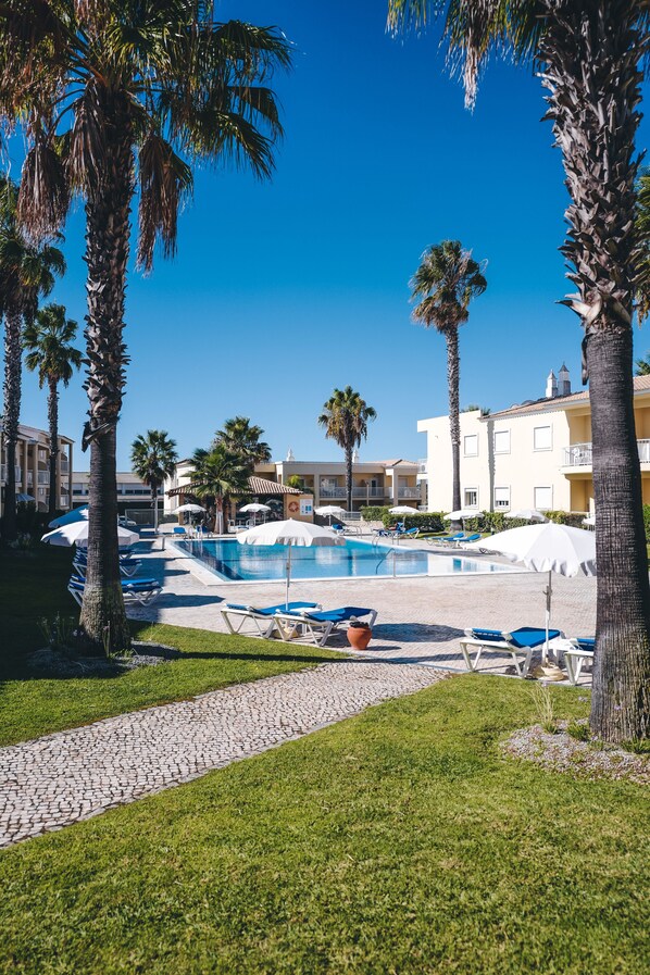 A pool area with lounge chairs and umbrellas, surrounded by palm trees and hotel buildings.