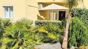 Um edifício amarelo com uma janela, um guarda-sol branco e várias plantas.