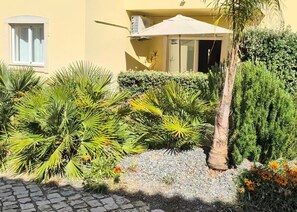 A yellow building with a window, a white umbrella, and various plants.