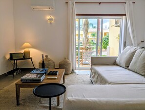 A room with a sofa, a coffee table with books, a lamp, and a view of a balcony with outdoor furniture.