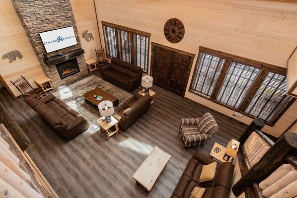 A spacious living room with a stone fireplace, a television mounted on the wall, and large windows with wooden shutters. - Private Indoor Pool & Basketball Court Retreat with Arcade Games, Sleeps 50, Free Tickets & Views (Branson)