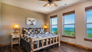 A bedroom with a large bed, two bedside tables, a ceiling fan, and large windows offering a view of greenery. - Private Indoor Pool & Basketball Court Retreat with Arcade Games, Sleeps 50, Free Tickets & Views (Branson)