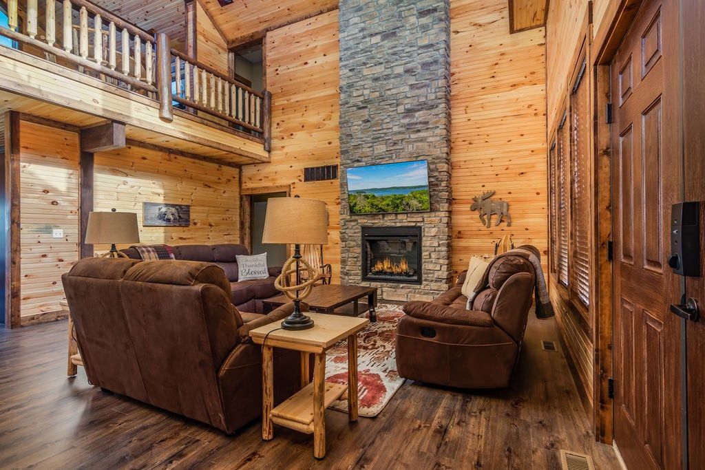 A log cabin interior with a fireplace, leather sofas, a dining table, and a balcony.