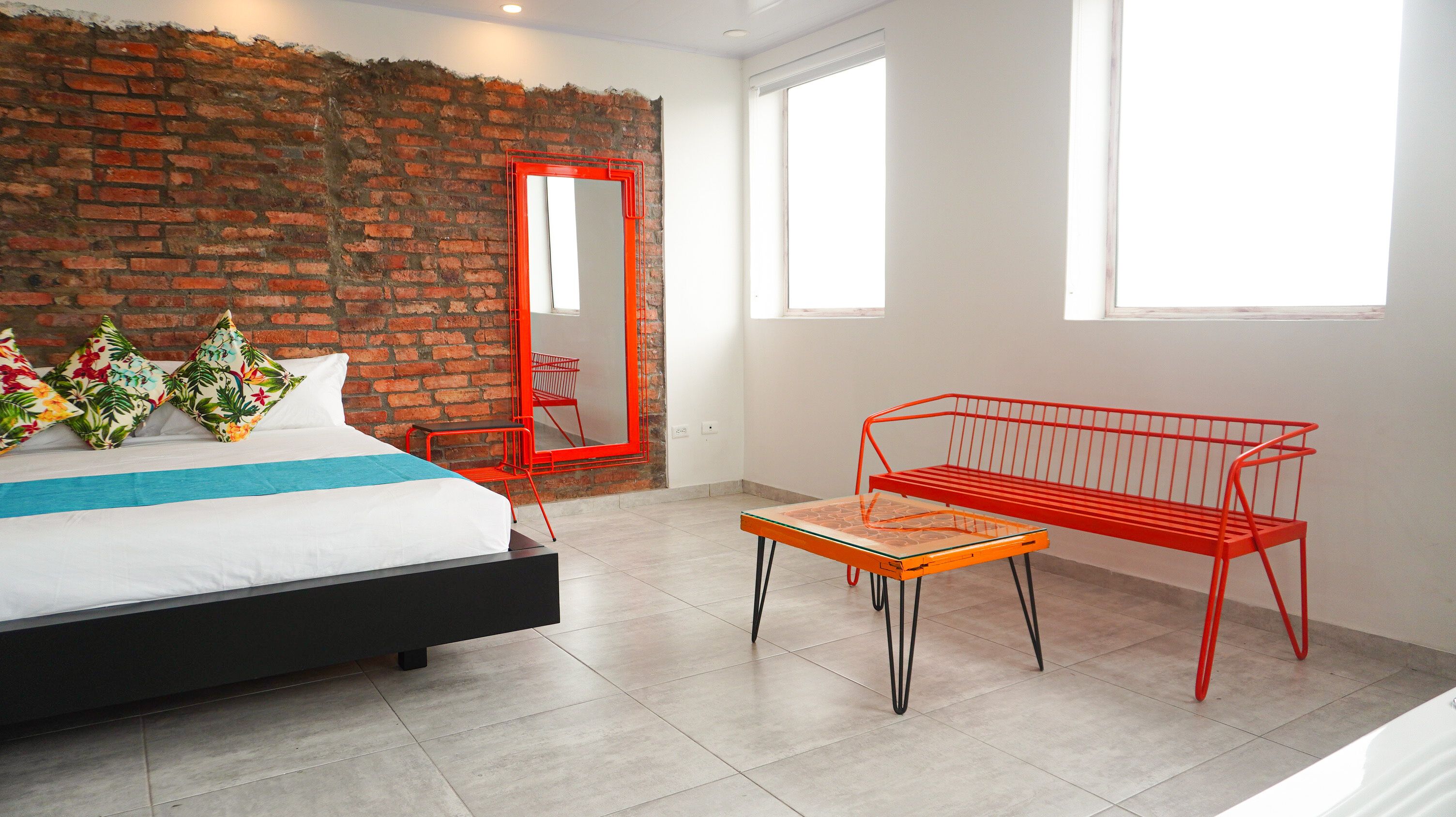 Une chambre moderne avec un mur en briques, un lit agrémenté d’oreillers à motifs floraux, un banc rouge et une grande fenêtre.