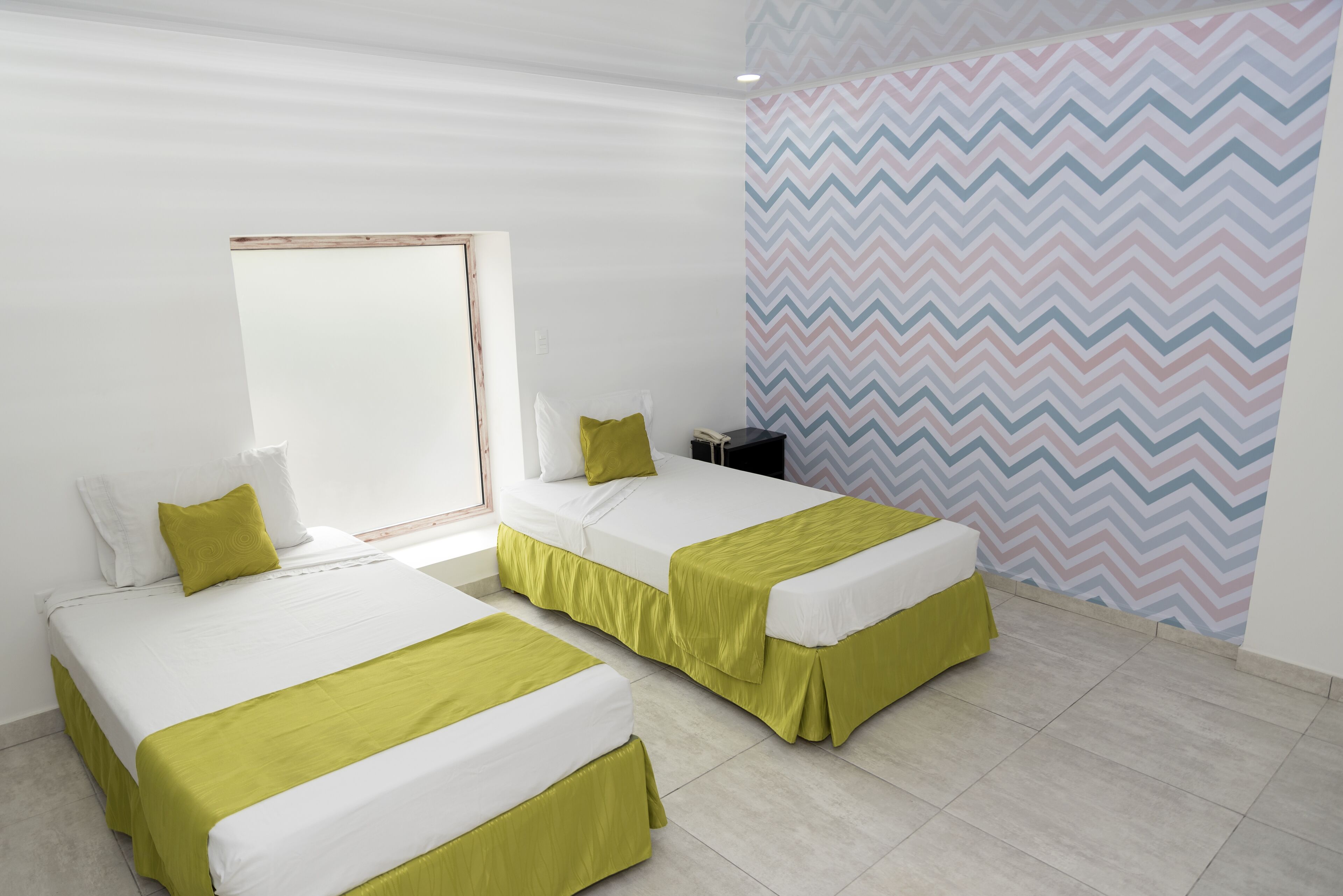 Habitación de hotel con dos camas, cada una con colcha verde y ropa de cama blanca. Se ve una ventana con marco de madera, una pared con diseño en zigzag y una mesita con un teléfono.