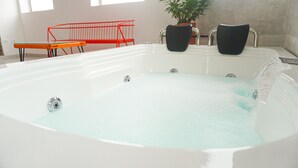 A large, white, freestanding bathtub with a clear water feature, a red chair, and a potted plant in a modern bathroom setting.