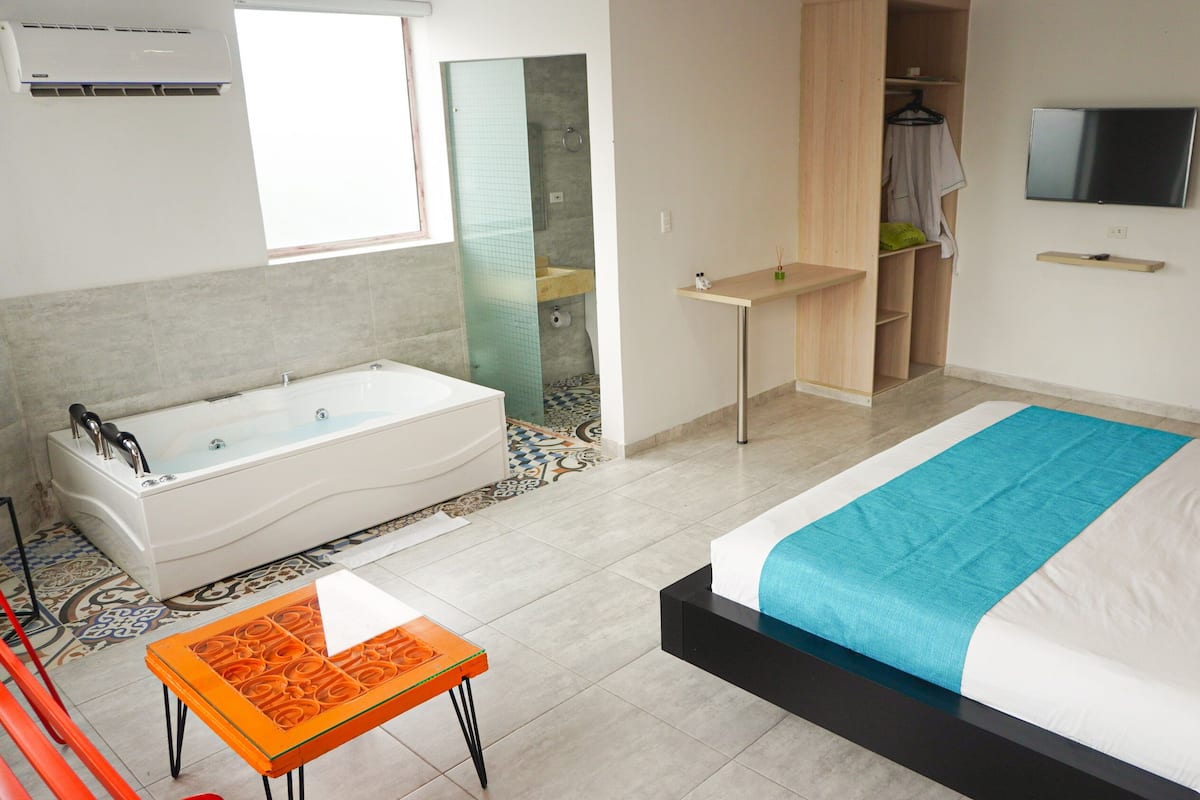 Un dormitorio moderno con una cama grande, un baño separado con bañera, un televisor montado en la pared y una mesita con una silla.