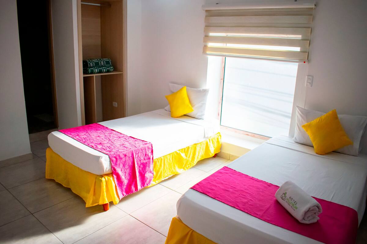 Habitación de hotel con dos camas, cada una con una colcha rosa y amarilla, y un ventanal con persianas.
