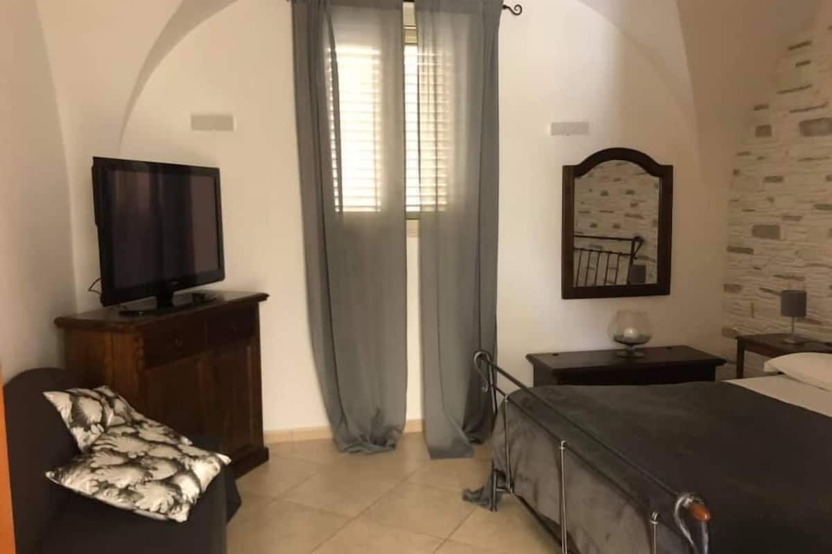 A bedroom with a bed, a TV on a wooden cabinet, a mirror, and a window with curtains.