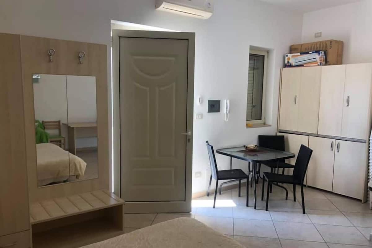 A room with a bed, a desk, a chair, a table, a wardrobe, and a door.