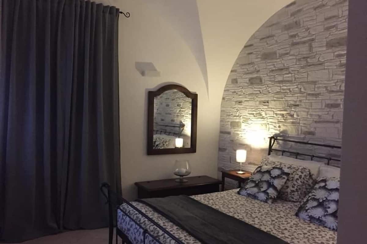 A bedroom with a stone wall, a bed with patterned bedding, a nightstand with a lamp, and a mirror on the wall.