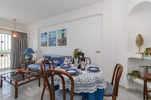 A dining area with a round table set for four, surrounded by wooden chairs. There is a sofa with blue cushions in the background, and a window with curtains to the left. - Club Atlantis 78 - One Bed (Puerto Colon)