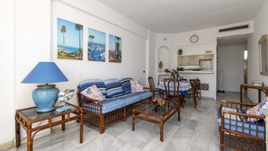Una sala de estar con sofá, sillas, una mesa de centro y un comedor con mesa y sillas. Se ven cuadros colgados en la pared y una zona de cocina al fondo.