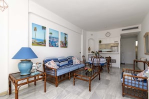 A living room with a sofa, chairs, a coffee table, and a dining area with a table and chairs. There are wall-mounted pictures and a kitchen area visible in the background. - Club Atlantis 78 - One Bed (Puerto Colon)