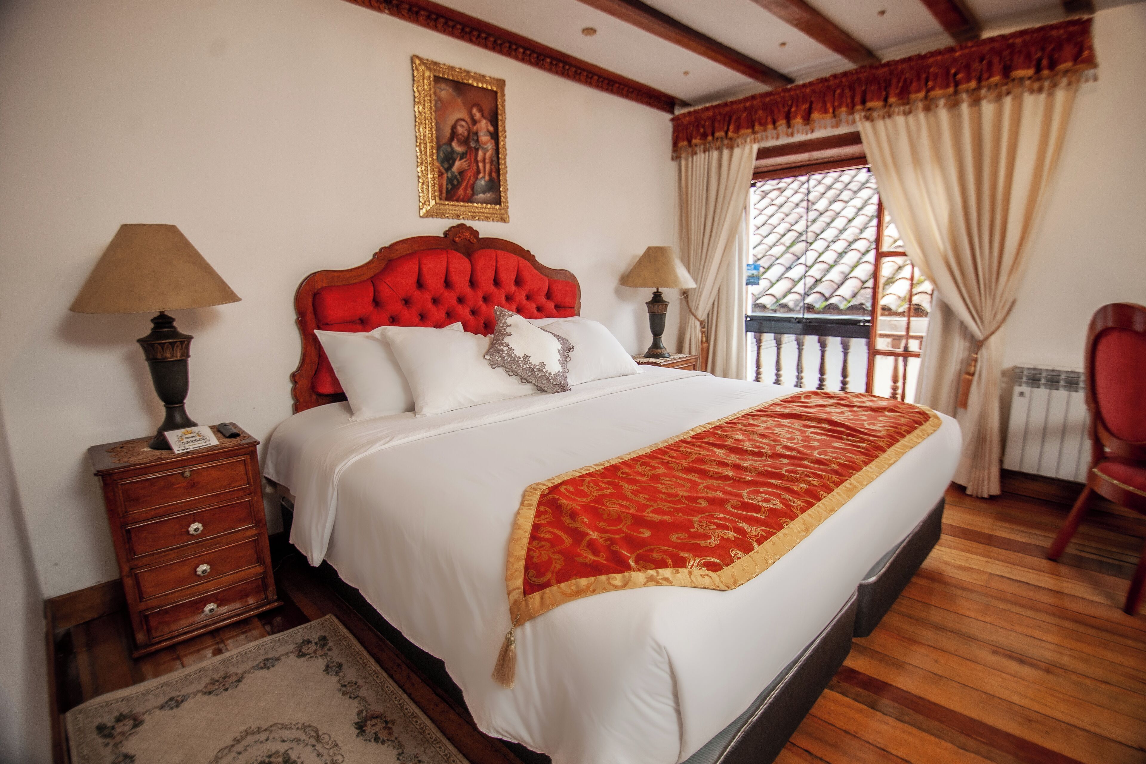 Un dormitorio con una cama grande, suelo de madera, una mesita de noche, una lámpara, una silla y una ventana con cortinas.