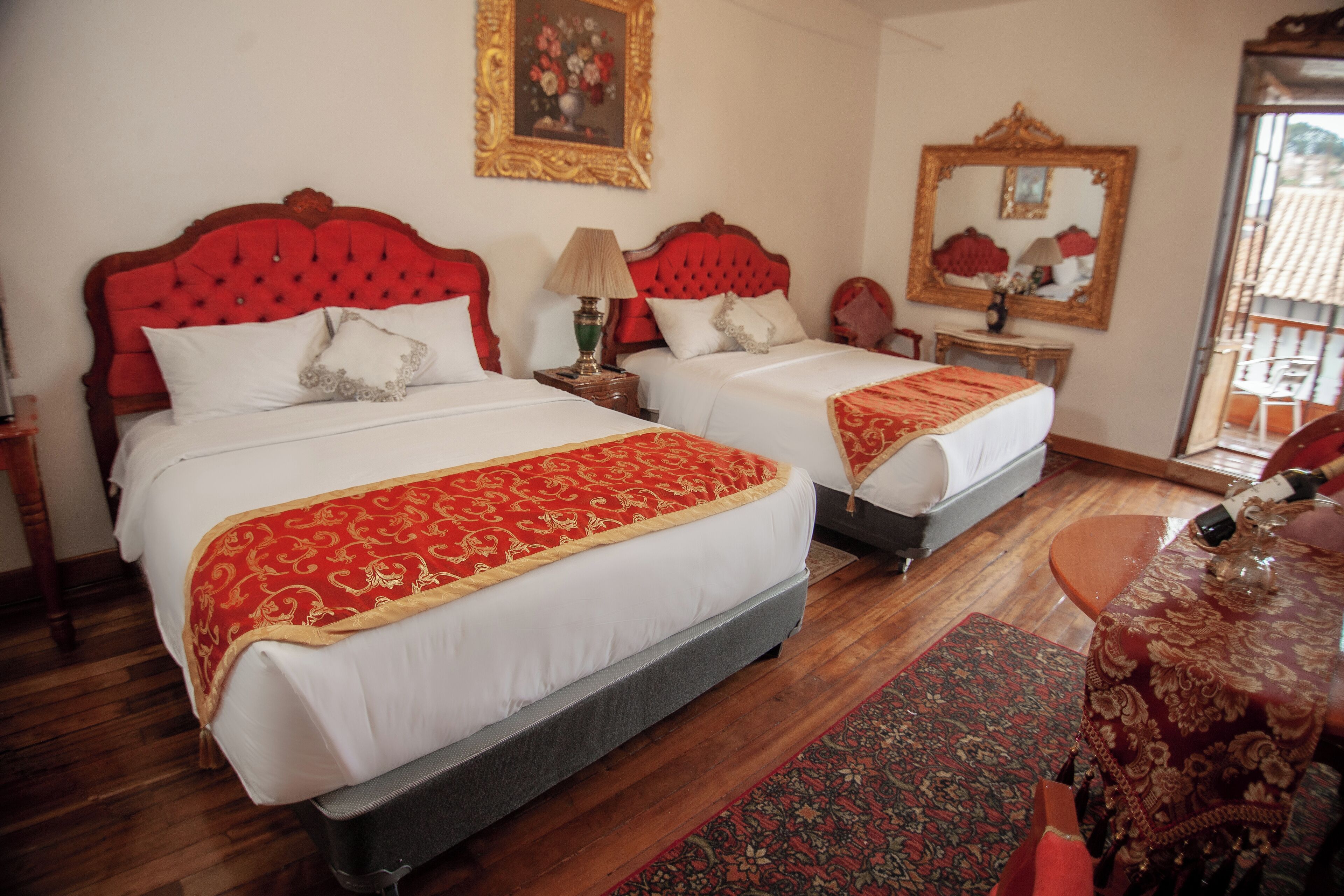 A hotel room with two beds, each with a red headboard and white bedding. There is a wooden floor, a small table with a lamp, and a mirror on the wall.