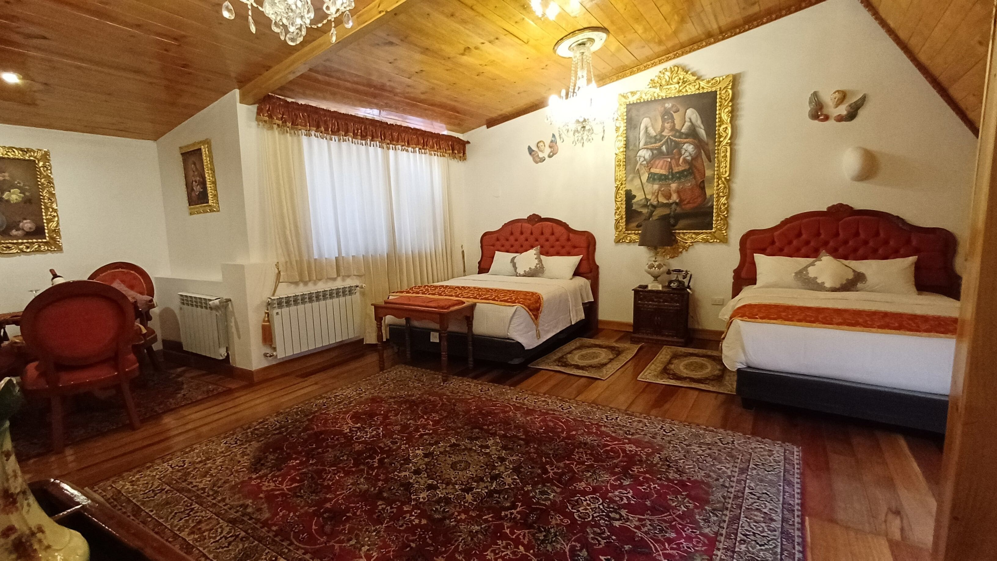 A hotel room with two beds, a red upholstered headboard, a large ornate chandelier, a red and gold patterned rug, and a painting of an angel on the wall.