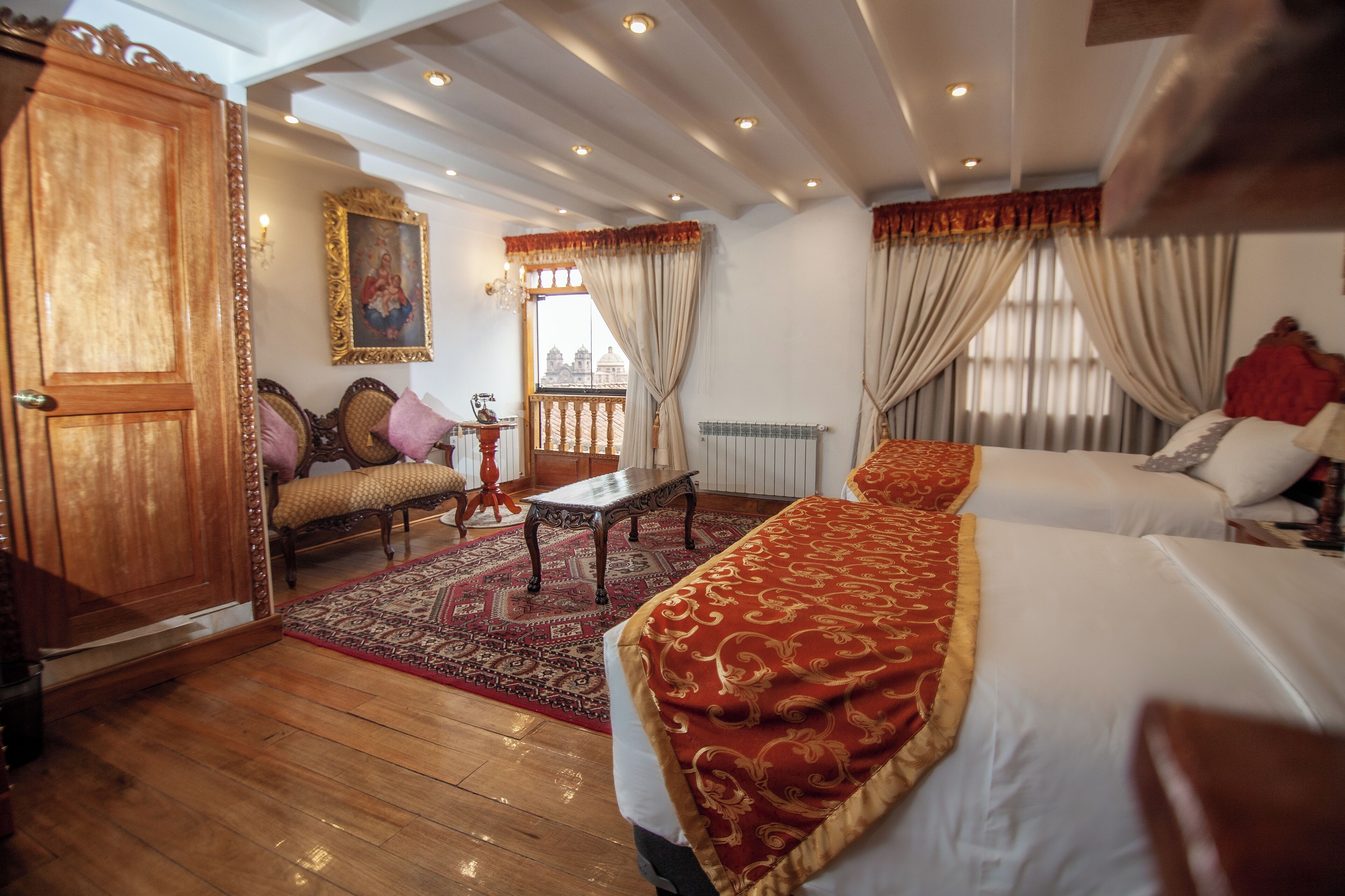 A hotel room with a large bed, a wooden wardrobe, a small table, a sofa, and a view of a cityscape through the window.
