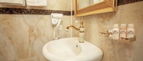 Un baño con un lavamanos blanco, grifo dorado y un espejo encima. Hay artículos de aseo en un estante y un dispensador de toallas en la pared.