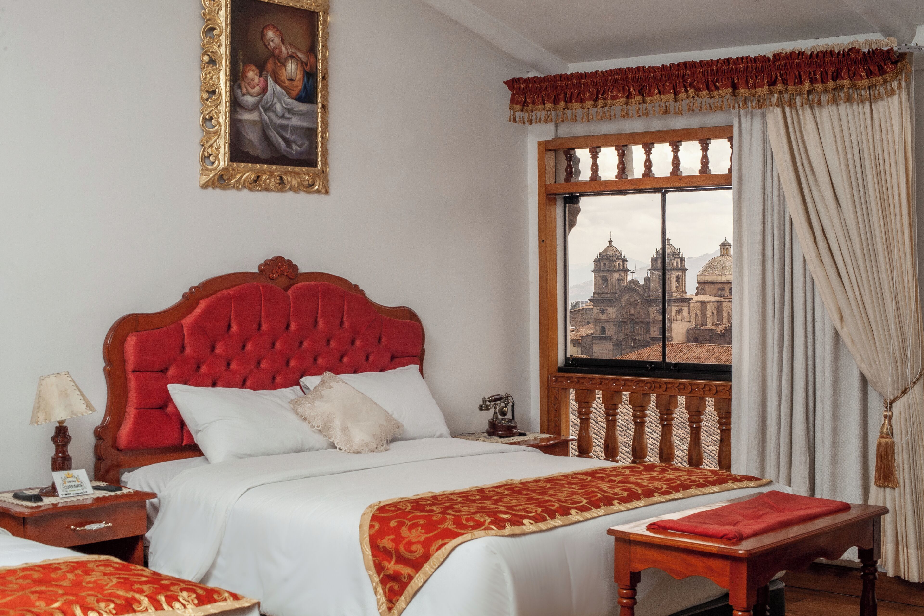 A hotel room with a large bed, a wooden nightstand, a table, and a window with a view of a cityscape.