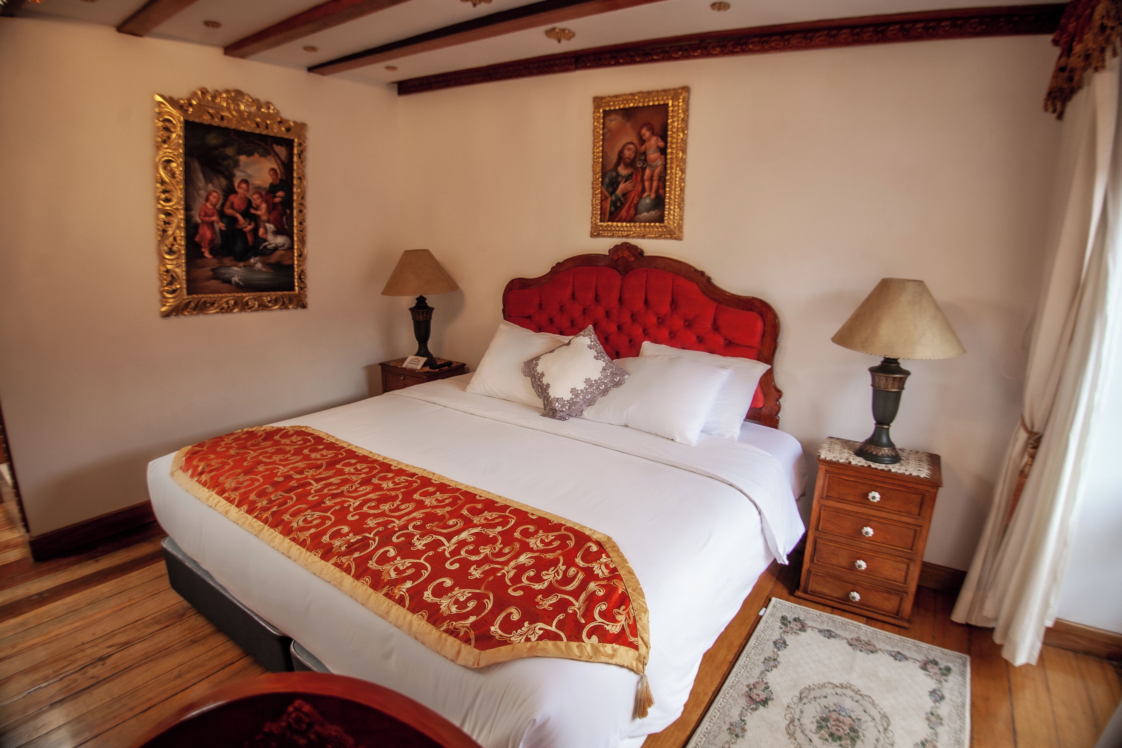 Uma cama bem arrumada com colcha vermelha e branca, duas mesas de cabeceira com abajures e duas obras de arte emolduradas nas paredes.