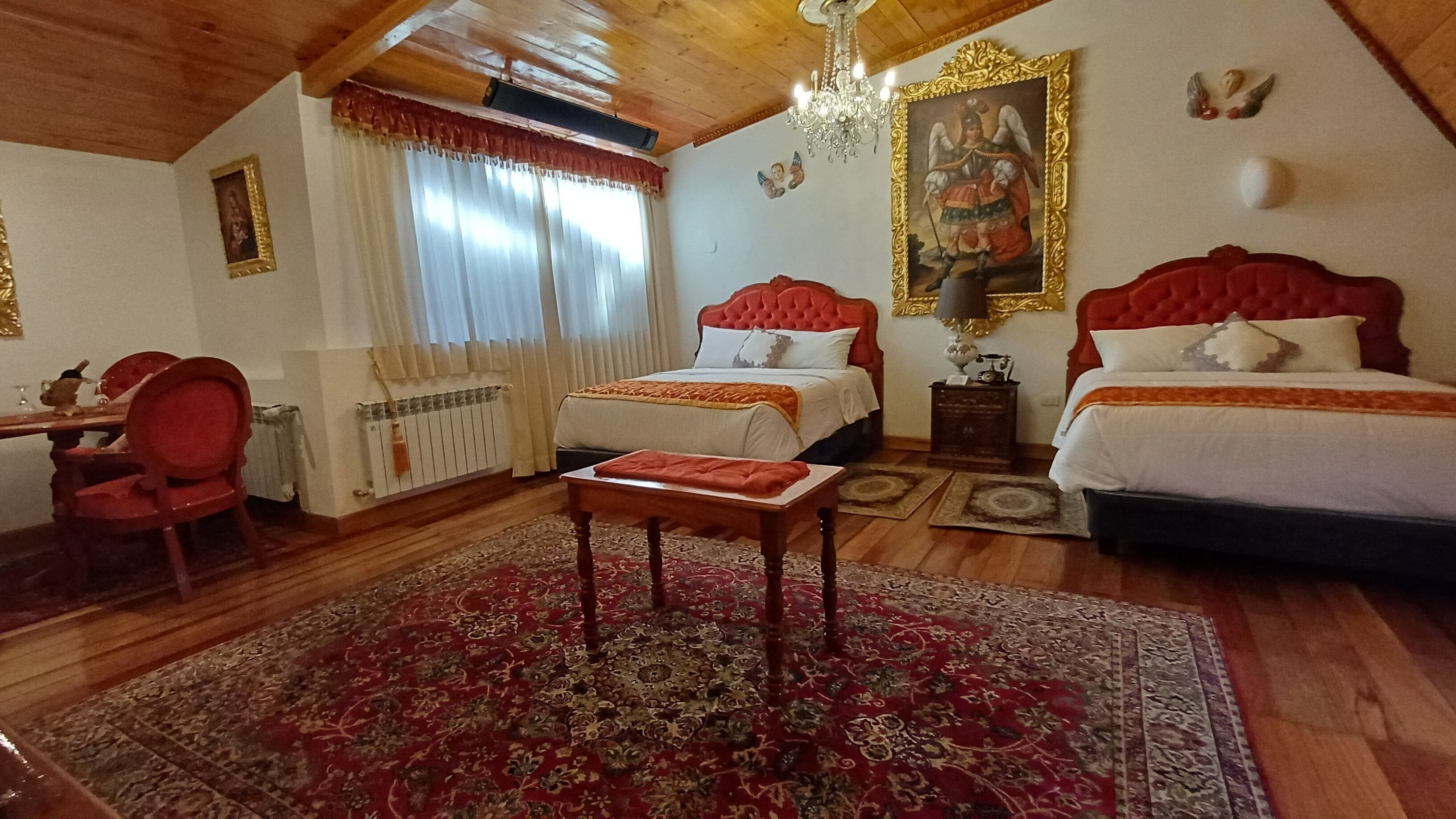 A hotel room with two beds, a wooden ceiling, a chandelier, a red and white patterned rug, and a painting on the wall.