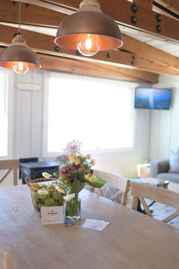 A wooden dining table with a vase of flowers and a basket of fruits. There are hanging pendant lights above the table. - Dillon Beach Resort (Dillon Beach)