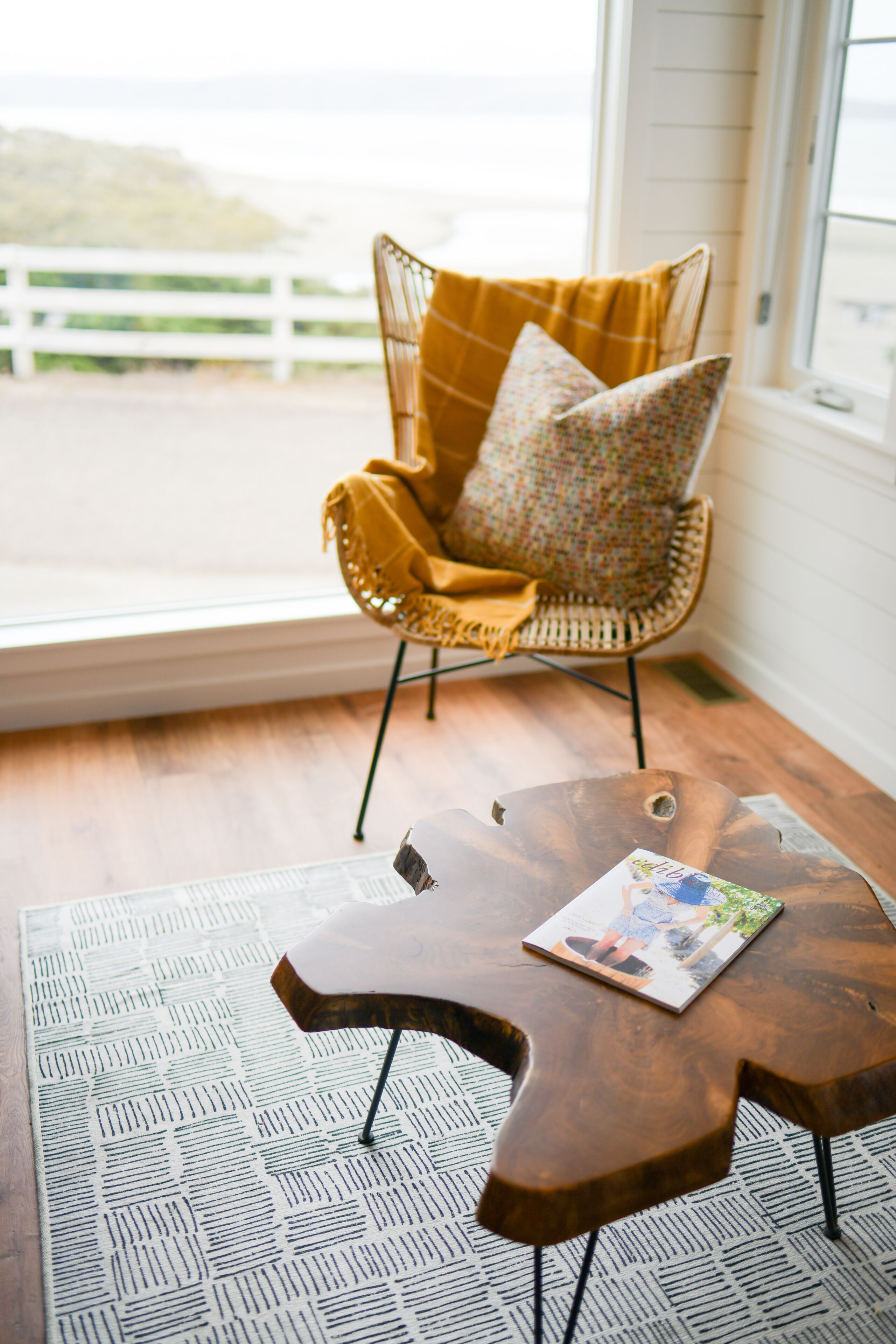 A room with a wicker chair with a yellow throw blanket, a wooden coffee table with a magazine, and a patterned rug.