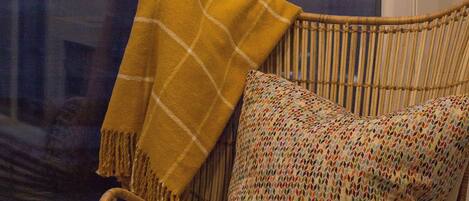 A wicker chair with a colorful pillow and a yellow throw blanket in front of a window with a sunset view.