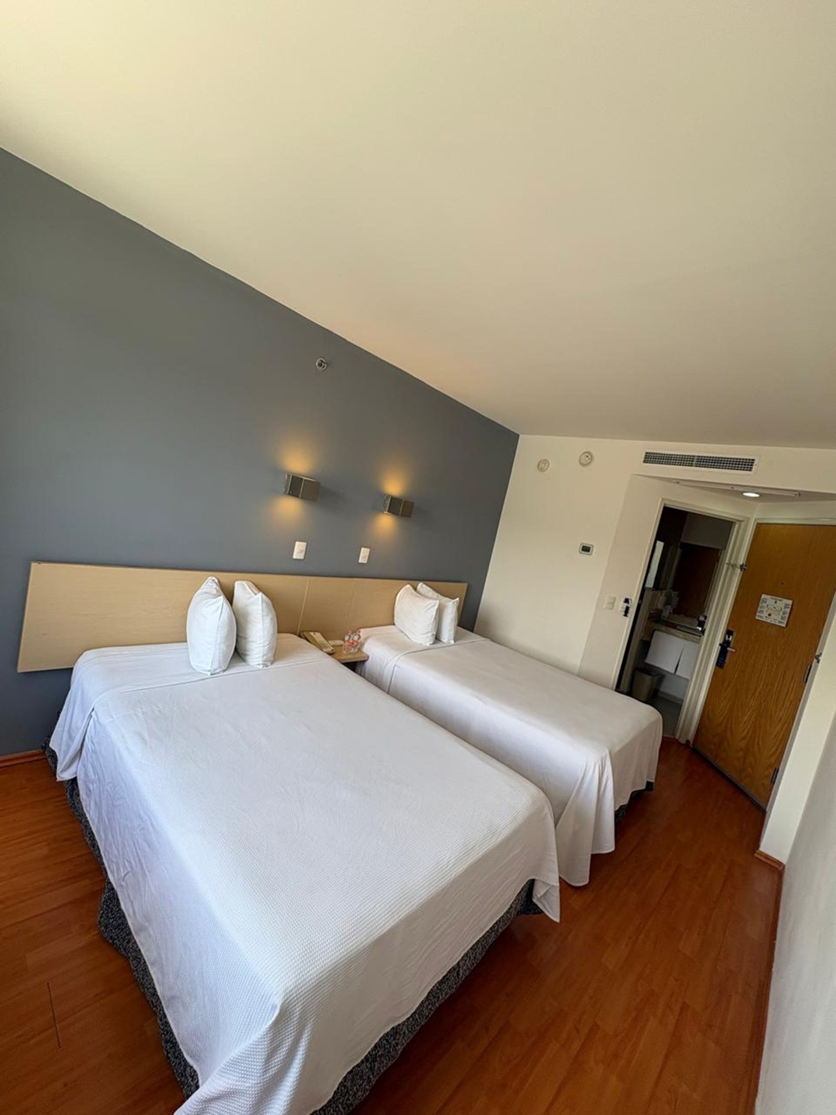 Une chambre d’hôtel avec deux lits, un plancher en bois, un mur d’accent de couleur grise et une porte donnant accès à la salle de bain.