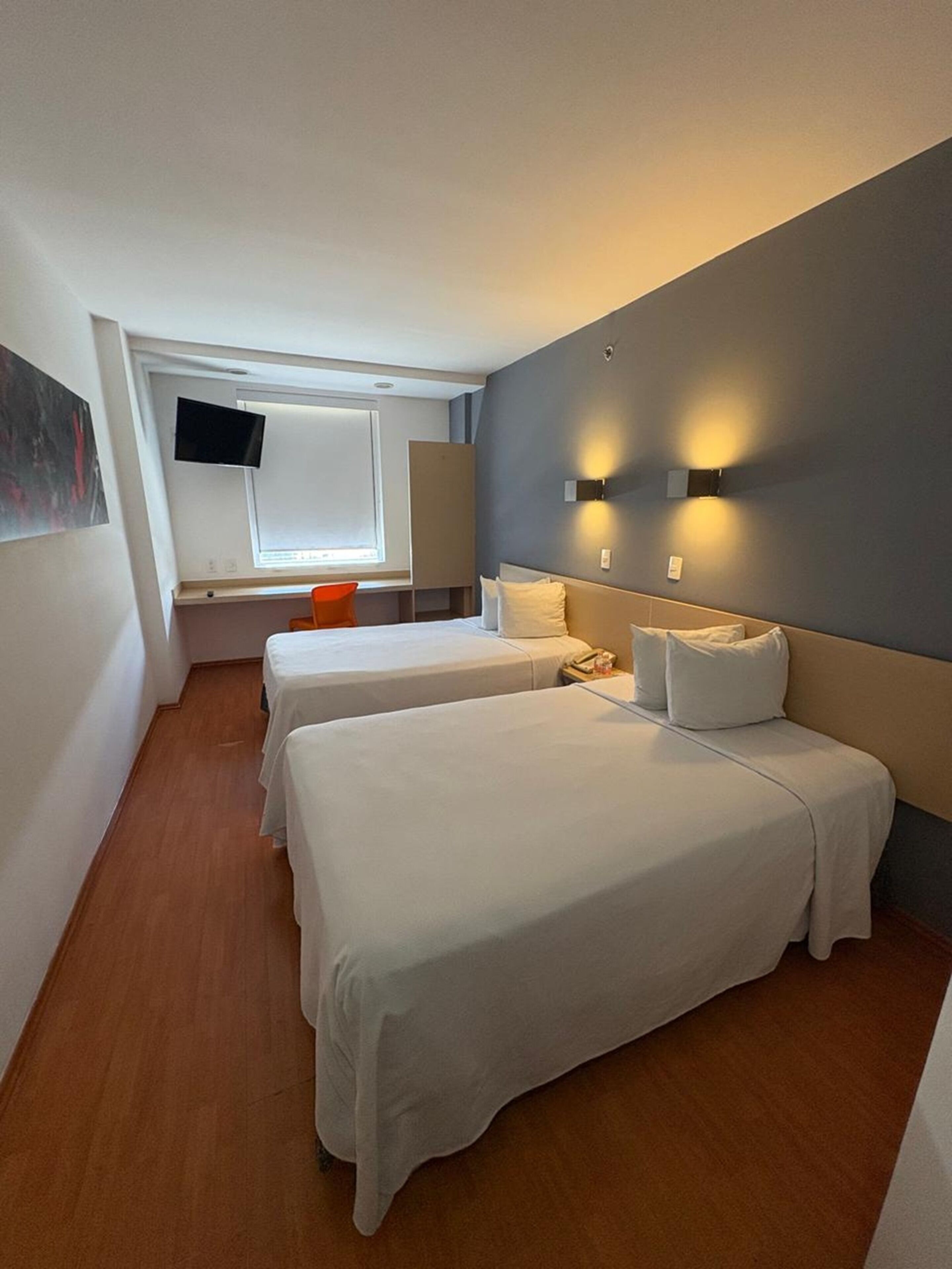 A hotel room with two beds, a TV, a window, and a wall-mounted light fixture.
