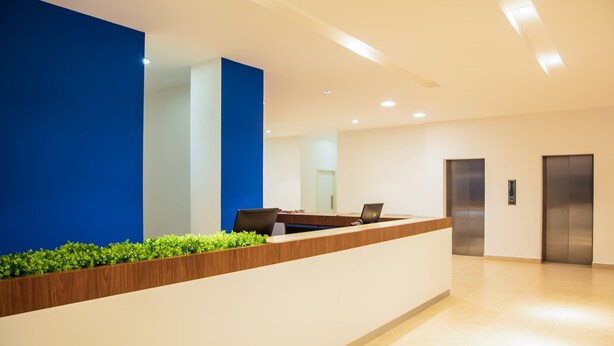 A reception desk with a blue accent wall, a wooden counter, and a laptop. There are two elevators in the background. - One Irapuato (Irapuato)