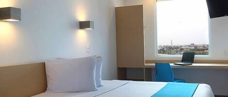 A hotel room with a bed, a desk with a laptop, a chair, and a window with a city view.