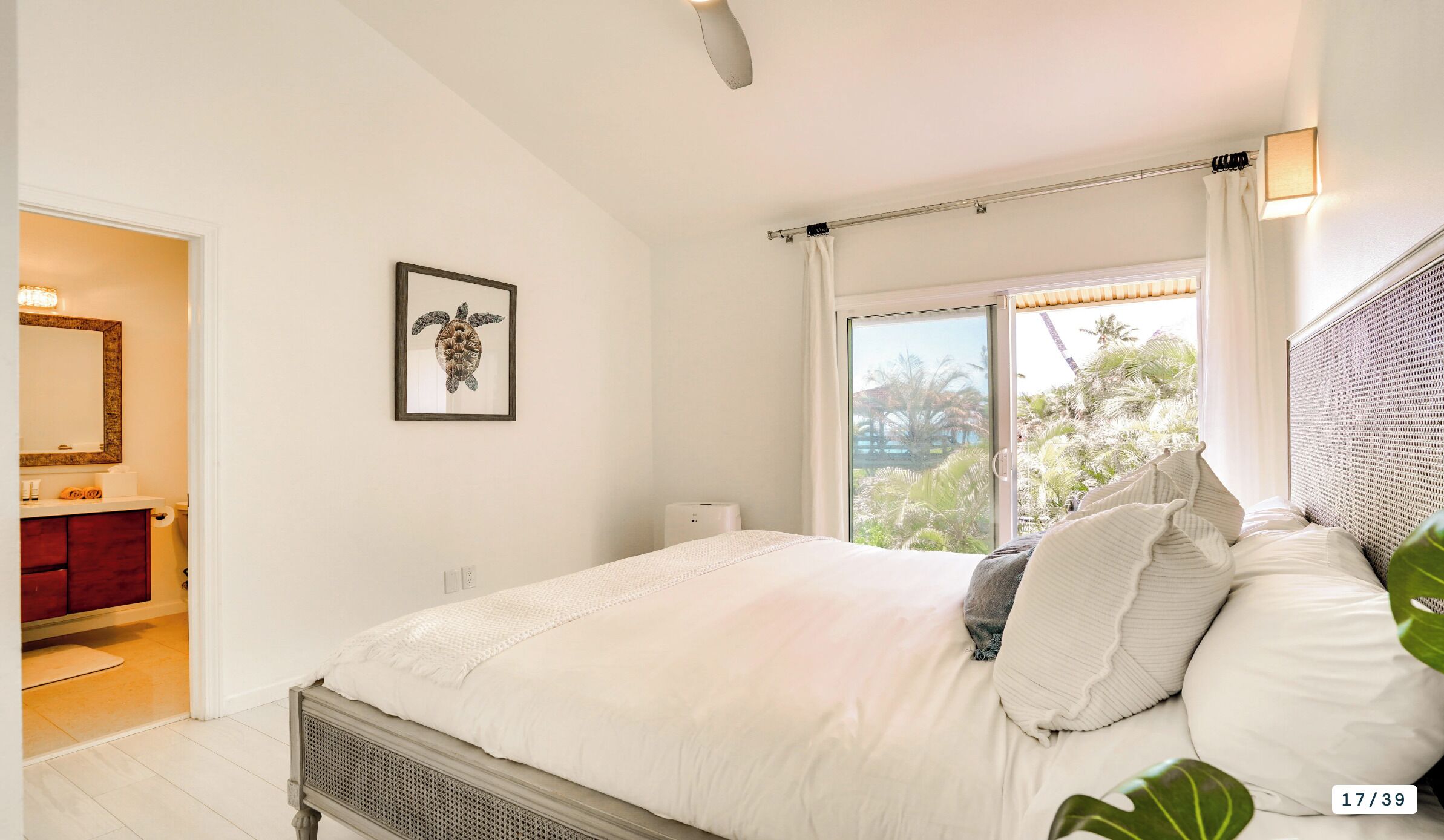 Une chambre avec un lit, une fenêtre donnant sur des palmiers, un tableau encadré au mur et une salle de bain avec un meuble rouge.