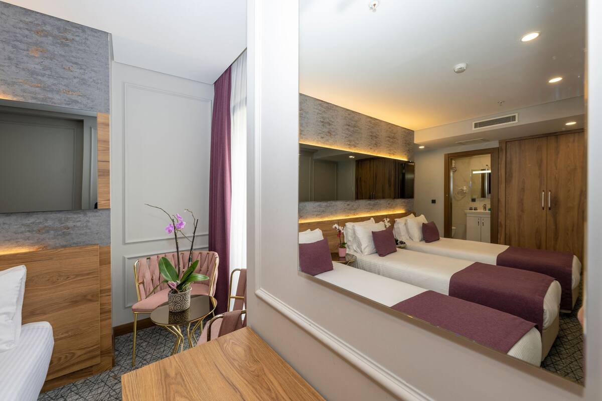 A hotel room with a bed, a desk, a chair, a small table, a potted plant, and a mirror.