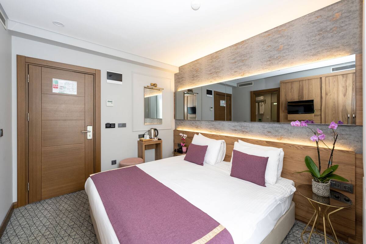 A modern hotel room with a large bed, a wooden wardrobe, a small desk with a chair, a mirror, and a potted orchid plant.