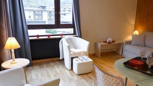 A modern hotel room with a view of buildings through large windows, a white sofa, a small table with a lamp, and a dining area with a glass table. - L'Aiguille du Midi by Interhome (Chamonix)