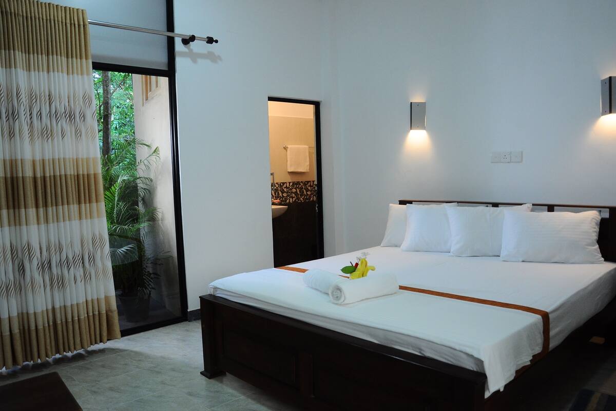 A hotel room with a large bed, a window with a view of greenery, a sliding door, and a wall-mounted light fixture.
