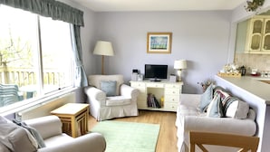 A cozy living room with a sofa, armchair, and a small wooden table. A television is on a stand, and there is a bookshelf with books and decorative items. - 14 Tweed Cottage (Jedburgh)