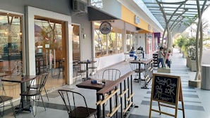 Un café al aire libre con mesas y sillas, techo de cristal y una pizarra publicitaria que anuncia una promoción.