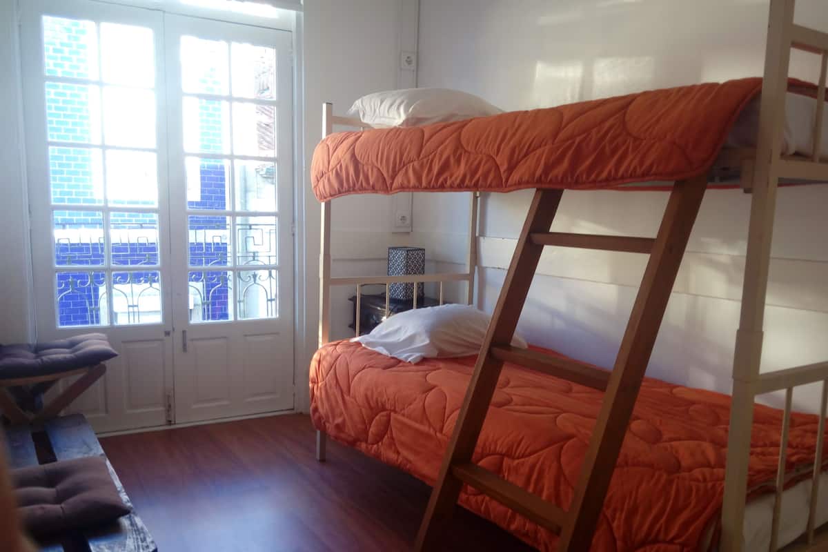 A bunk bed with an orange quilt, a wooden ladder, a white pillow, and a small table with a cushion.