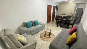 A living room with a grey sofa, a grey armchair, and a coffee table. There is a dining area with a table and chairs. - Family bungalow near the sea (Centro)