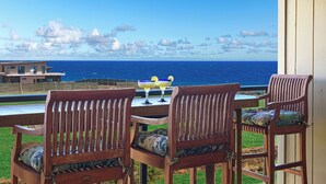 Une terrasse en bois avec deux chaises et une table, d’où l’on peut admirer un océan bleu limpide et un bâtiment situé au loin.