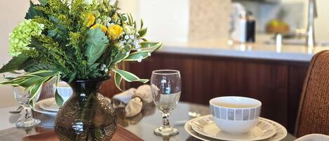 Une table à manger ornée d’un vase en verre rempli de fleurs, d’un verre à vin et d’un bol posé sur un sous-verre en bois.