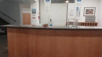 A reception desk with a wooden finish, a bottle of hand sanitizer, and a glass of water. There are signs on the wall, including one with a blue logo and another with a red emergency exit sign.