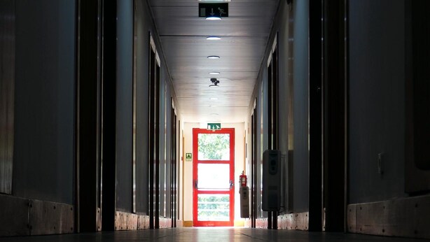 A long, narrow hallway with a red door at the end, flanked by columns, and a ceiling with recessed lighting.