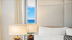 Hotellihuone, jossa on suuri ikkuna, valkoisilla lakanoiden peitetty sänky, yöpöytälamppu ja kuvioitu pääty.
