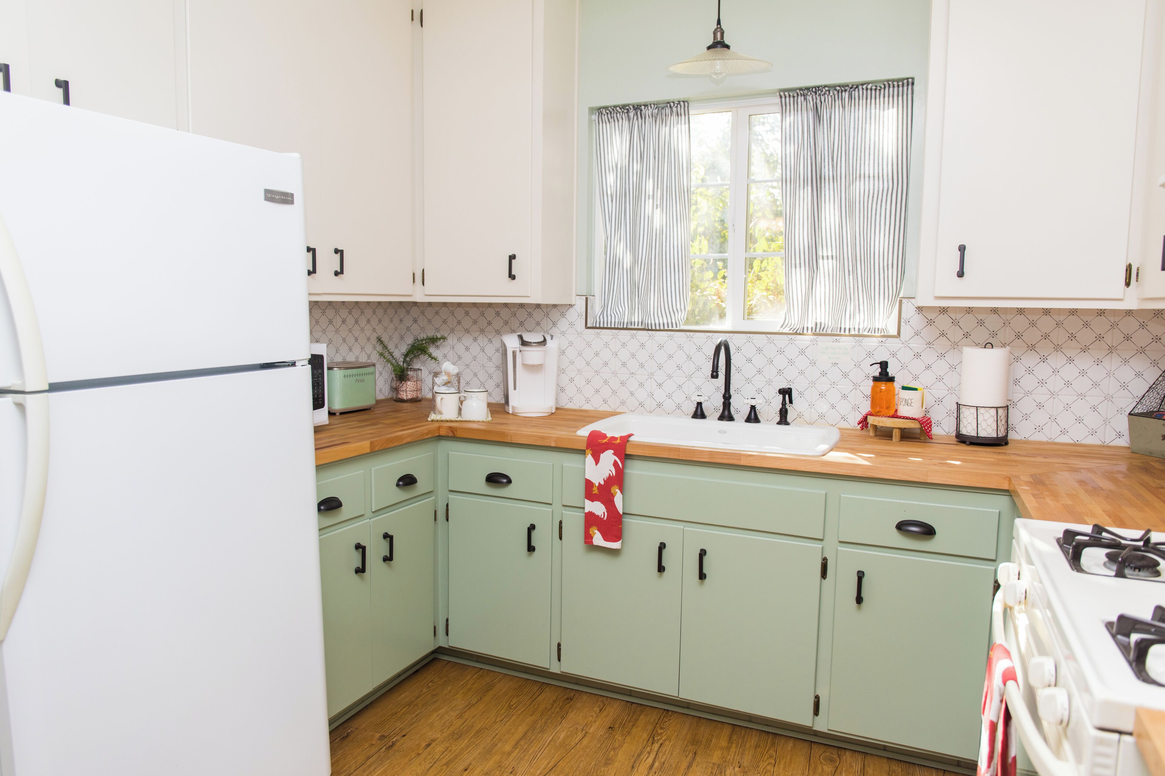 Una cuina amb armaris verdosos, un refrigerador blanc, un taulell de fusta, una barra, una finestra amb cortines i una cuina a gas.