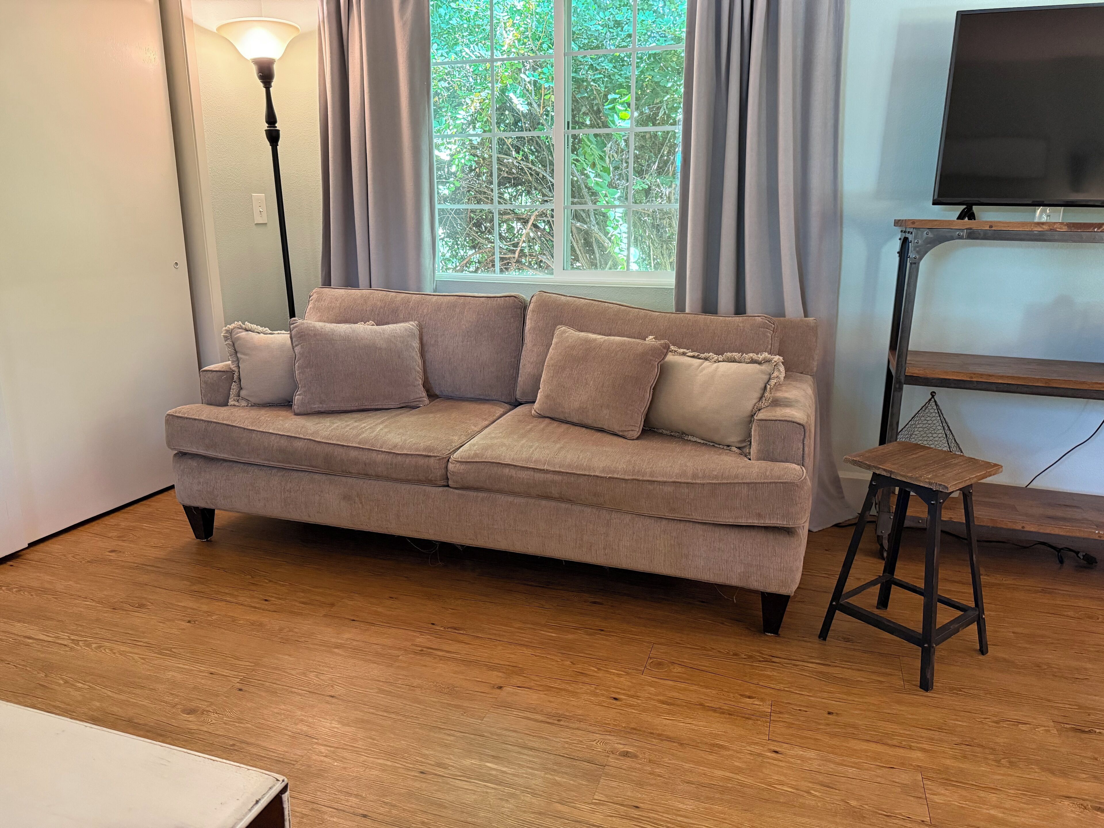 En beige sofa med tre puder, en lille træskammel, et fjernsyn på et stativ og et vindue med gardiner.