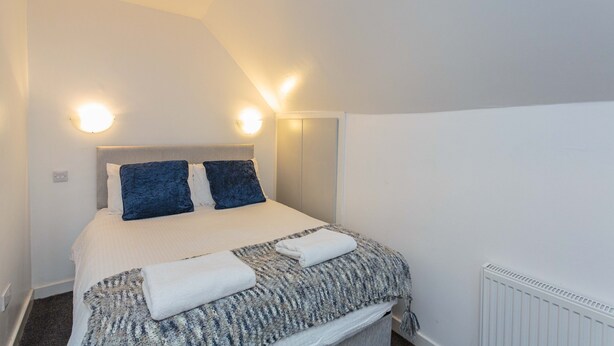 A bedroom with a bed, two pillows, a blanket, and a radiator. - Cherry Property - Blueberry Suite (Blackpool)